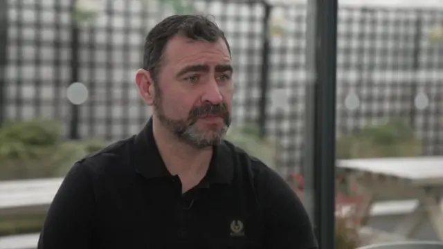 أندي ريد يجلس مرتدياً قميصاً أسود اللون، بوجه خالٍ من التعابير، في صورة من الكتفين إلى أعلى. هو رجل في منتصف العمر ذو شعر داكن ولحية داكنة، مع بعض الشعيرات الرمادية الخفيفة. يجلس أمام نافذة، وخلفه تظهر بعض المقاعد في حديقة.