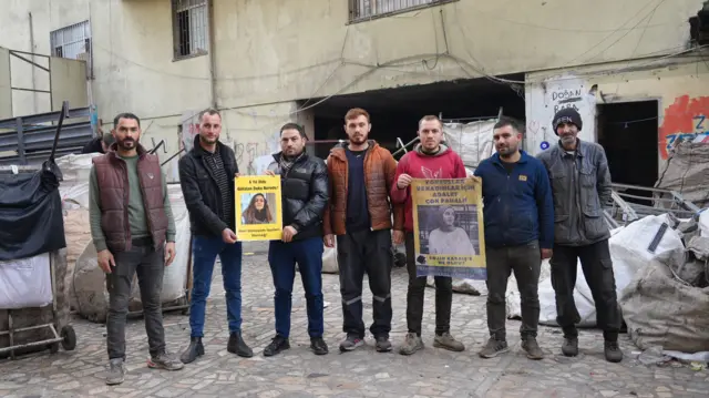 Geri dönüşüm işçileri ellerinde Gülistan Doku ve Rojin Kabaiş'e destek afişleriyle deponun avlusunda poz veriyor. Arkalarında ve yanlarında çuvallar gözüküyor.