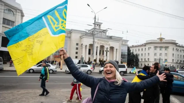 Радостная женщина с украинским флагом, на фоне обнимаются люди.