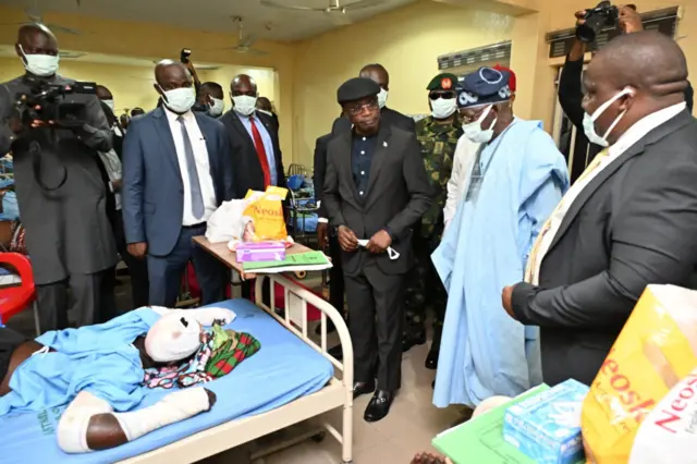 President Bola Tinubu weat sky blue clothe and navy blue cap stand beside Benue State Govnor, Rev Father Hyacinth Alia wey wear all balck suit as dem visit victims of di Benue killings for hospital