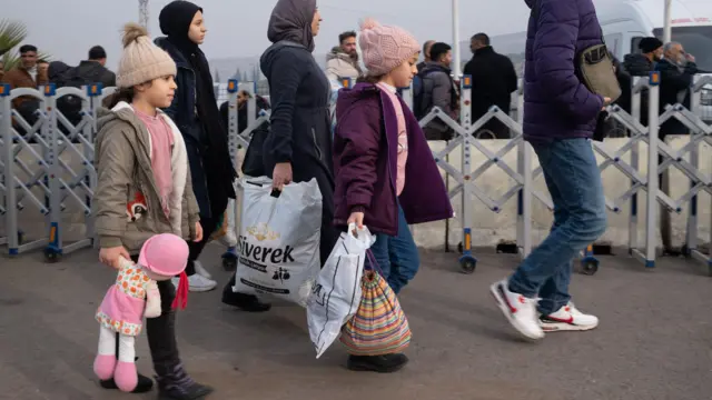 Duas crianças e três adultos caminham ao ladounibet 365 liveuma cerca, carregando sacolas e brinquedos