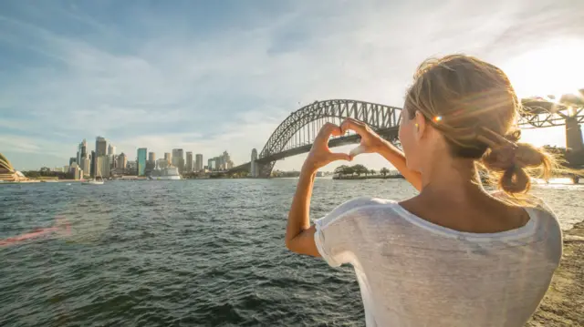 Mulher faz um coração com as mãos diante da costa de Sydney.