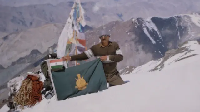 बर्फ से ढकी पर्वत चोटी पर सुरक्षात्मक वस्त्र पहने एक व्यक्ति ने एक हरा झंडा पकड़ रखा है, जिसके ऊपर एक छोटा भारतीय ध्वज है. 