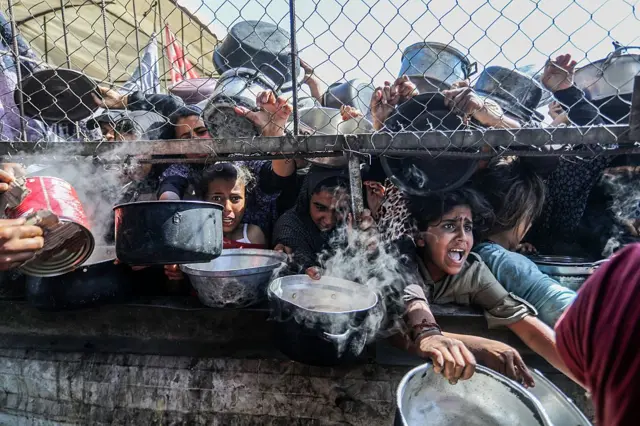 Gazze'deki bir yardım dağıtımı sırasında bağıran çocuklar, çitlerin arkasından boş tencerelerini uzatıyor.