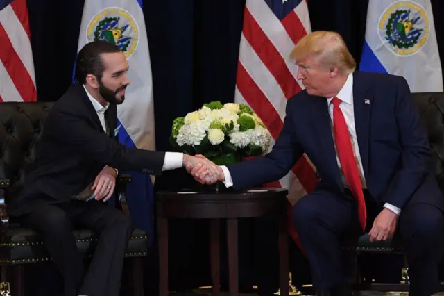 El presidente de El Salvador, Nayib Bukele y el presidente de Estados Unidos, Donald Trump sentados, se estrechan la mano con las banderas de sus países al fondo. 