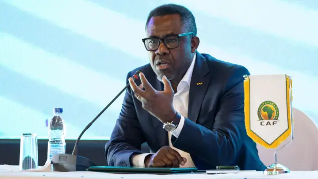 Le secrétaire général de la Caf, Veron Mosengo-Omba, parle à une table supérieure pour une conférence de presse, avec un logo de la Caf sur un support à côté de lui.