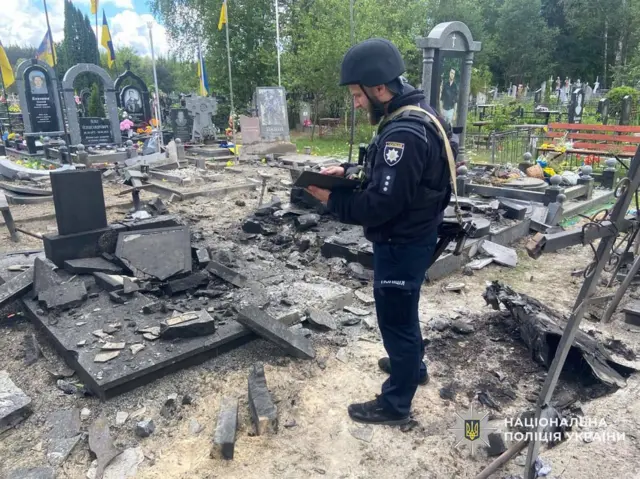 На кладовищі видно зруйновані надгробки та розкидані уламки каменю, що свідчать про сильне руйнування. Поруч стоїть працівник у формі та шоломі, який щось занотовує, фіксуючи наслідки пошкоджень.