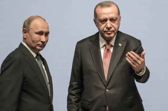 putin ve erdoğan bir sahnede, üstlerinde takım elbiselerle yan yana duruyor.