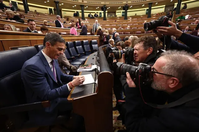 Pedro Sánchez sentado en su escaño en el Congreso de los Diputados mientras un grupo de fotógrafos lo retratan. 