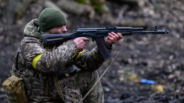 A soldier dressed in camo and a green hat crouches while aiming an assault rifle into his the distance