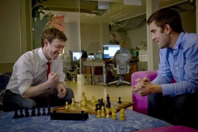 Two people - one of them Mark Zuckerberg - are playing chess. The chess board is perched on a blue, patterned foot stool. Mark Zuckerberg is playing with white pieces, his opponent black. Mr Zuckerberg is clenching his right fist and smiling.