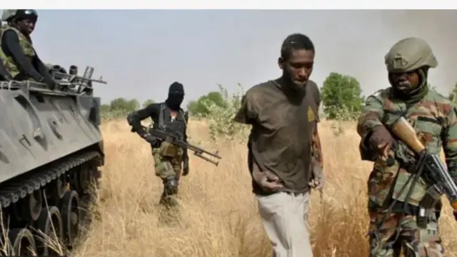 Ṣọja kan n bọ lẹyin pẹlu ibọn, agbesunmọmi ti wọn mu wa laaarin, ṣọja kan naa wa niwaju rẹ, o n tọka ọna ti yoo gba fun un. Ọkọ ologun wa ninu aginju ọhun pẹlu, ṣoja kan jokoo lorii rẹ, o n wo agbesunmọmi ti wọn n mu lọ.