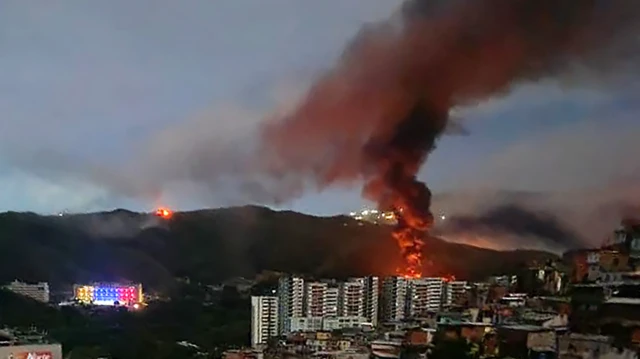 Una nube de humo a lo lejos en medio de la ciudad de Caracas, donde se ven viviendas y edificios y una montaña