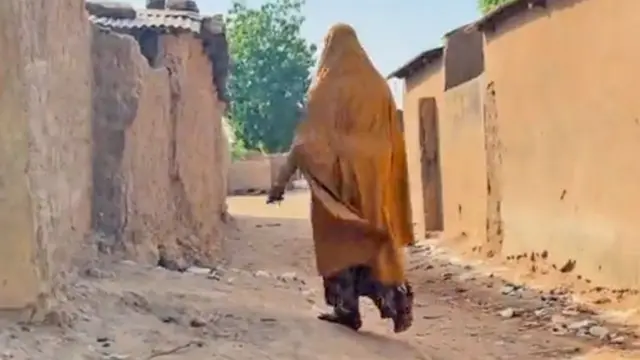 Une femme vêtue d'une robe couleur moutarde, vue de dos, marchant dans une  rue de Nukuru bordée de bâtiments aux murs de terre.