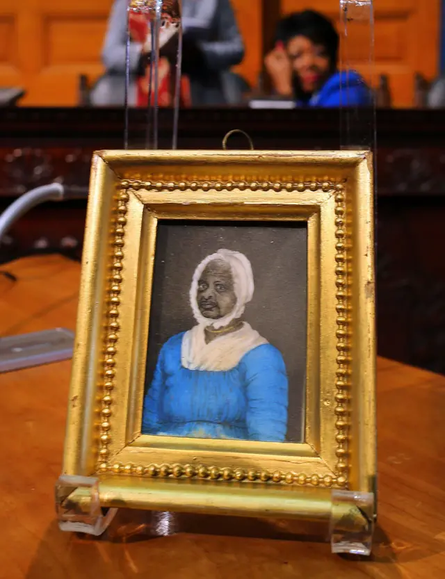 Pequeño retrato enmarcado de una persona con vestido azul y cofia blanca, exhibido sobre una mesa.