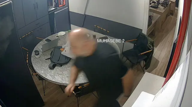 CCTV görüntülerinde bir odada koşarak koşan bir kişi görülüyor. Yanında bir masa ve dolaplar var. 