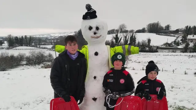 Tres niños pequeños junto a un muñeco de nieve que hicieron. Todos llevan guantes, gorros y abrigos, y se agarran a trineos rojos.