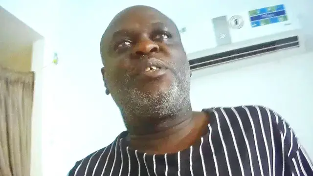 Charles Mwesigwa afise umushatsi mukemuke n'umumerera w'ubwanwa bw'imvi yambaye agapira k'imirongo ishinze mu mabara y'ubururu n'iryera