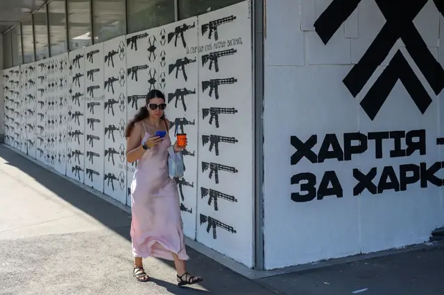 Молода жінка в світлій сукні й сонцезахисних окулярах іде тротуаром, дивиться в телефон і тримає напій. Позаду – довга стіна з повторюваними чорними зображеннями зброї та написом "Хартія за Харків".