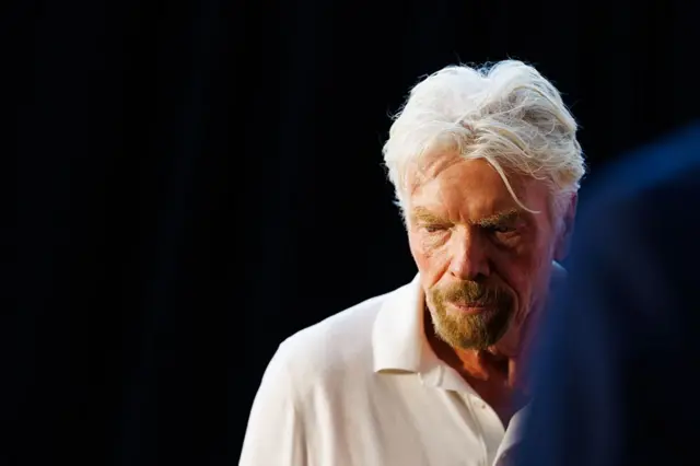 Richard Branson é um homem de cabelos grisalhos. Ele usa uma blusa branca e está olhando para o chão. 