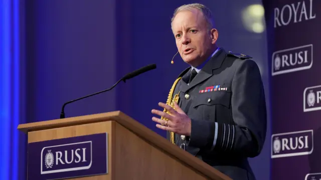 Mężczyzna w mundurze wojskowym stoi przy mównicy i wygłasza przemówienie. W tle i na mównicy widoczne jest logo RUSI.