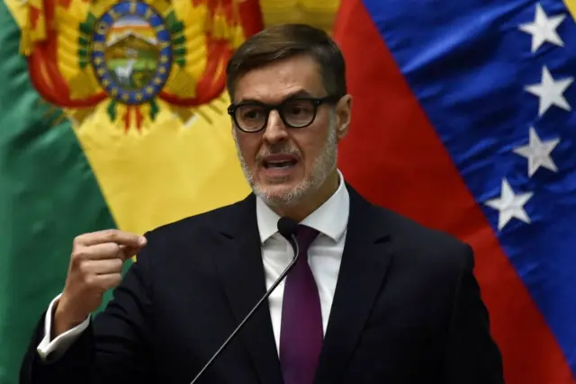 Félix Plasencia en primer plano con chaquea y corbata hablando ante un micrófono con las banderas de Bolivia y Venezuela de fondo. 