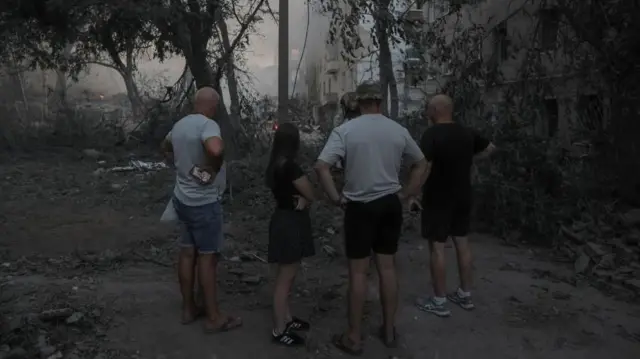 Três homens de shorts e camiseta e uma jovem com roupas leves, de pé em um jardim, observando a fumaça que sai dos destroços causados por um ataque em Kramatorsk, atribuído ao exército russo.