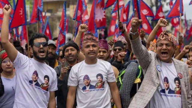 राजा ज्ञानेन्द्र र रानी कोमलको तस्बिर अङ्कित टीशर्ट लगाएका राजतन्त्रवादीहरू, पृष्ठभूमिमा नेपालका झण्डाहरू छन्
