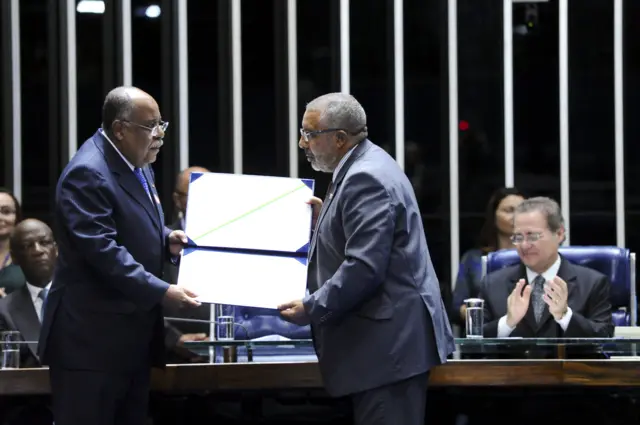 O ministro Benedito Gonçalves e o senador Paulo Paim no Plenário do Senado. Ambos são homens negros idososjogo ouro betterno. Paim estende um diploma comemorativo a Benedito