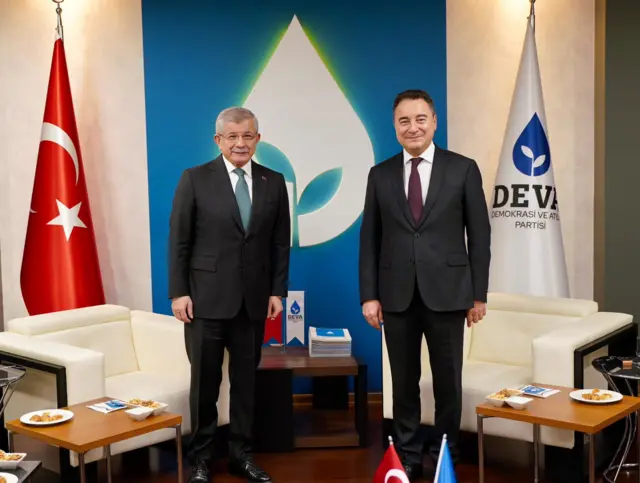 İki takım elbiseli erkek yan yana poz veriyor. İkisi de gülümsüyor. Arkada Türk bayrağı ve DEVA Partisi logosu var.