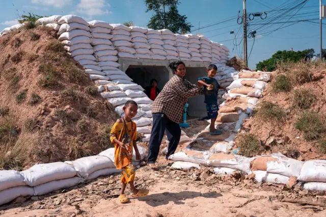 ประชาชนในพื้นที่ จังหวัดสระแก้ว ที่ไม่อพยพออกนอกพื้นที่ นั่งหลบในบังเกอร์เพื่อความปลอดภัยโดยมี ชรบ. วางกำลังเฝ้าหมู่บ้าน หลังช่วงเช้าที่ผ่านมามีรายงานว่ากัมพูชาได้ยิงจรวด BM-21 ลงบ้านเรือนประชาชนในพื้นที่ดังกล่าว ส่งผลให้บ้านเรือนประชาชนเสียหาย 2 หลัง โดยไม่มีผู้บาดเจ็บหรือเสียชีวิต เนื่องจากผู้นำชุมชนได้สั่งอพยพชาวบ้านไปยังศูนย์พักพิงและบังเกอร์ทันเวลา เมื่อ 9 ธ.ค 2568