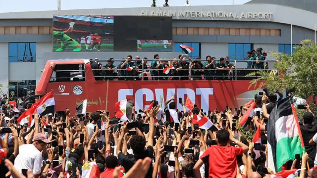 لاعبو المنتخب المصري يلوحون بأيديهم من على متن حافلة مكشوفة، وسط حشود من المشجعين الذين تجمعوا لاستقبالهم في مطار العلمين الدولي بمدينة العلمين الساحلية الشمالية في 10 يوليو/تموظ عام 2026. ويظهر مشجعون يلوحون للاعبين ويحملون أعلام مصرية، ويحاول بعضهم التقاط صور بهواتفهم المحمولة.
