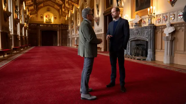 Леви и принц Уильям стоят в одном из залов Виндзорского зала. На полу красный ковер, за спиной принца камин.