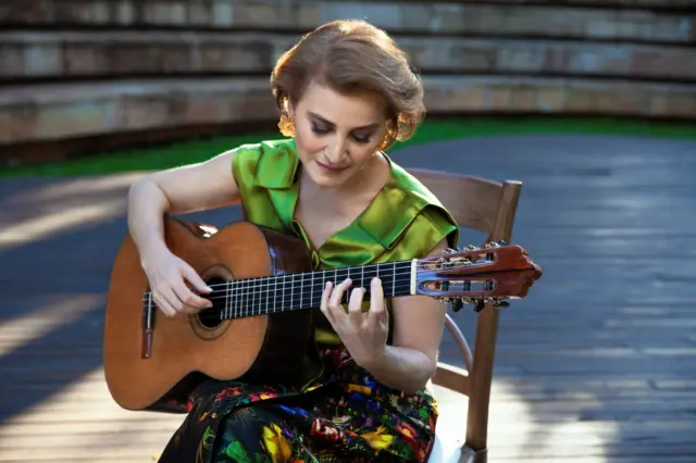 Berta Rojas toca su guitarra. Está sentada en una silla de madera. 