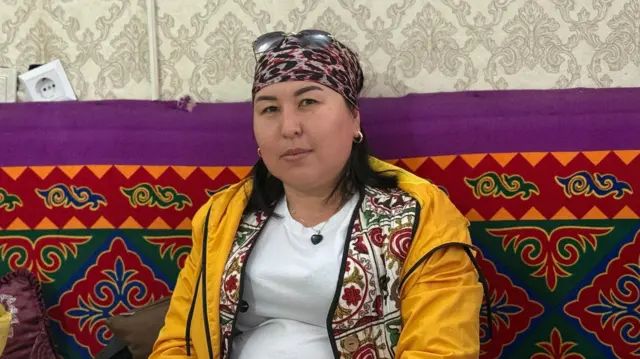 Kırgız motiflerinin olduğu bir minderin üzerinde oturan, sarı hırka giyen bir kadın.