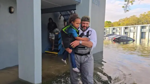 
برایان استرن در سال ۲۰۲۴ در منطقه سیل‌زده‌ای در تامپا، فلوریدا، در جریان طوفان میلتون، کودکی خردسال را در آغوش گرفته است. آب تا زانوهایش رسیده و ماشین‌ها در پس‌زمینه در آب فرو رفته‌اند.