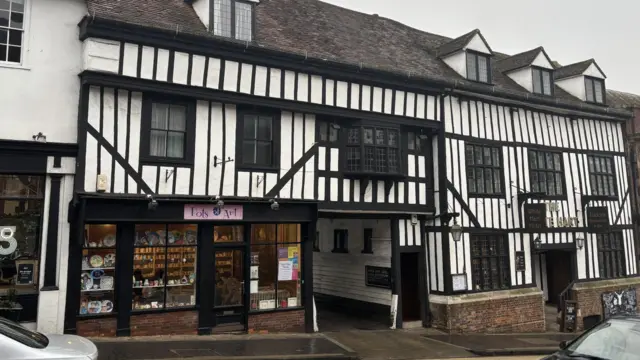 Exterior de un edificio de estilo Tudor, con marcos de madera blancos y negros y pequeñas ventanas. Hay un callejón en el centro, un hotel a la derecha y una tienda a la izquierda.