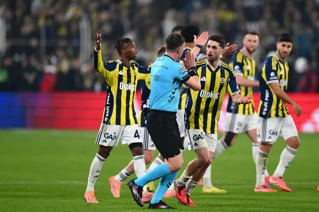 Fenerbahçeli oyuncular mavi renkli üniformalı hakeme itiraz ediyor.