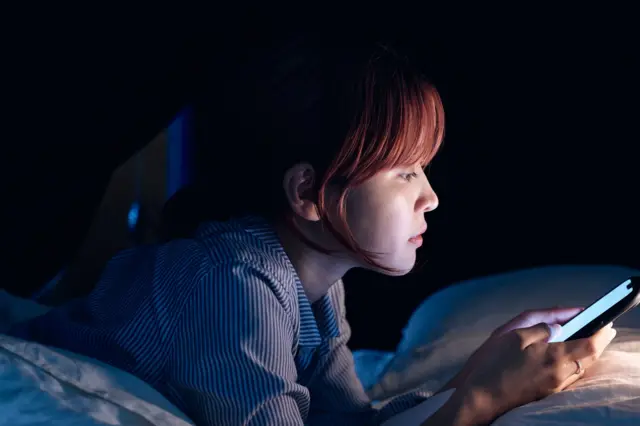 Una joven en su cama, en una habitación oscura, con su teléfono.