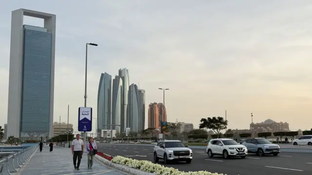 городской пейзаж Абу-Даби: на заднем плане высотки, на переднем видна автодорога, вдоль нее по тротуарам идут люди в легкой одежде европейского вида