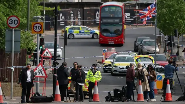 A road scene showing members of the media work by a police cordon, in front of police cars and a parked bus, while a British flag flies overhead.
