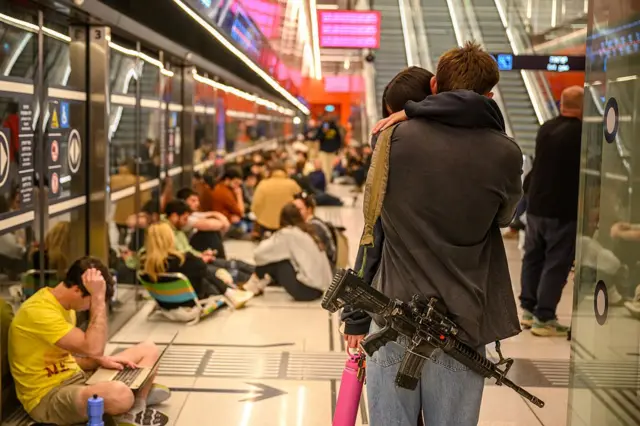 Silahlı bir delikanlı sevgilisine sarılmış. Metro kapılarının önünde yerde oturan insanlar var. 