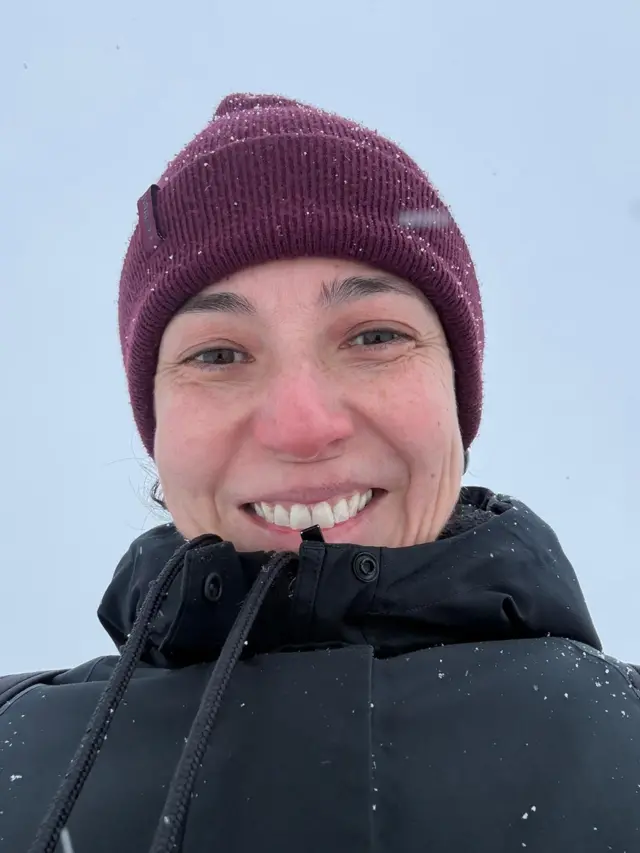 A designer brasileira Jane Vita sorri para a câmera em um ambiente nevado, com flocos de neve visíveis sobre o gorro de lã roxo e o casaco preto fechado até o queixo; o rosto está levemente avermelhado pelo frio, com pequenas sardas e marcas naturais de expressão, enquanto o fundo é um céu cinza uniforme.