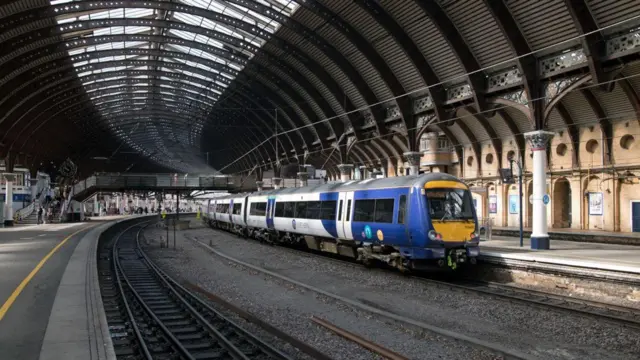 Picture of the inside of York railway station, there is a Northern train pulling in at platform 14. You can see a bridge going over the tracks with people walking along it.