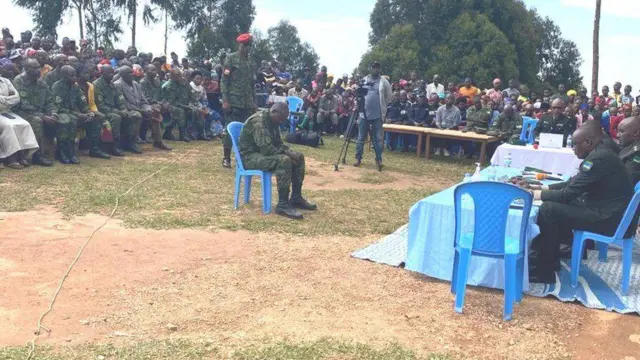 Sergeant Gervais Minani yicaye ku ntebe ya plastike, yambaye imyambaro y'igisirikare cy'u Rwanda. Inyuma ye hahagaze umusirikare wambaye ingofero itukura umurinze, imbere ye hari abacamanza ba gisirikare. Bakikijwe n'imbaga irimo abaturage n'abasirikare, bamwe barahagaze abandi baricaye. Ifoto yo ku wa mbere, ku itariki ya 9 Ukuboza (12) mu mwaka wa 2024