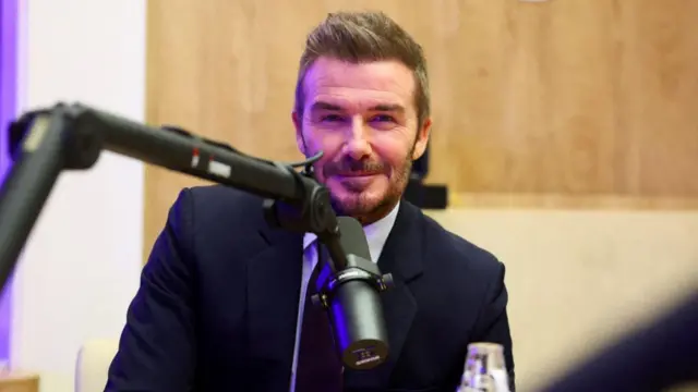 Üzerinde takım elbise olan David Beckham tebessüm ederek poz veriyor. Önünde bir mikrofon var.