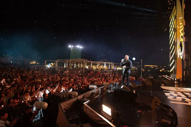 Эмин Агаларов поет на сцене под открытым звездным небом. На фото видны зрители, многие из которых снимают его на мобильные телефоны