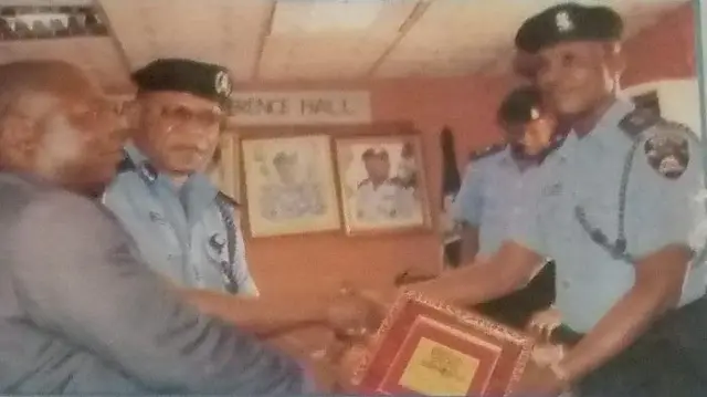 Adegbehin Adetarami wọ asọ ọlọpaa, o n gba ami ẹyẹ lọwọ ọkunrin kan to wọ asọ kootu dudu, to si n bọ ọ lọwọ nigba to n gbe ami ẹyẹ naa fun. Bakan naa, awọn osisẹ ọlọpaa meji miran, ti wọn wọ asọ ọlọpaa naa wa nibẹ pẹlu.