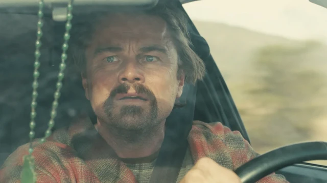 Leonardo DiCaprio caracterizado como su personaje, vestido con una camisa de cuatros, mira alertado al costado mientras sujeta el volante en una escena de "Una batalla tras otra".
