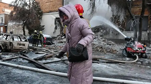 Ukrayna'nın Kiev kentinde, 12 Ocak 2026'da Rus insansız hava aracı saldırısının ardından yıkılan bir binada itfaiyeciler yangını söndürürken bir kadın yürüyor.